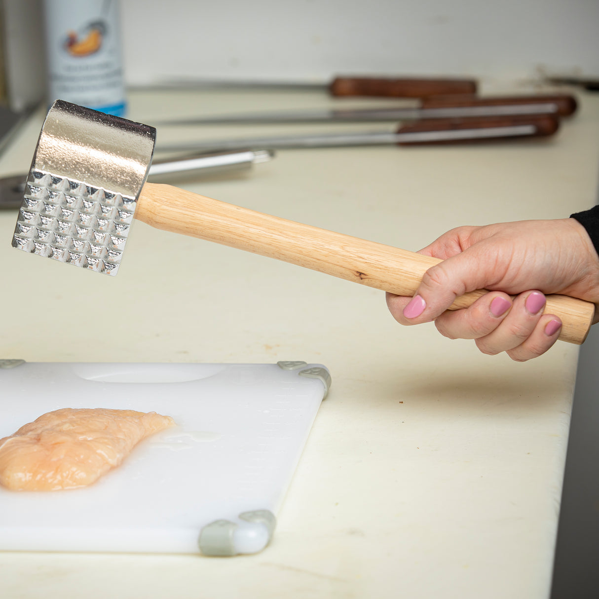 A hand with pink nail polish grips a wood handle meat tenderizer over a raw chicken breast on a white cutting board. Several knives with wooden handles are lined up in the background on a cream-colored countertop.