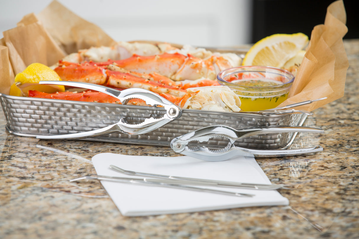 A tray of crab legs with lemon wedges and dipping butter sits on a granite counter. Chrome plated Lobster Crack double jaw crackers, seafood forks, and napkins are arranged in front, suggesting a seafood meal ready to eat.