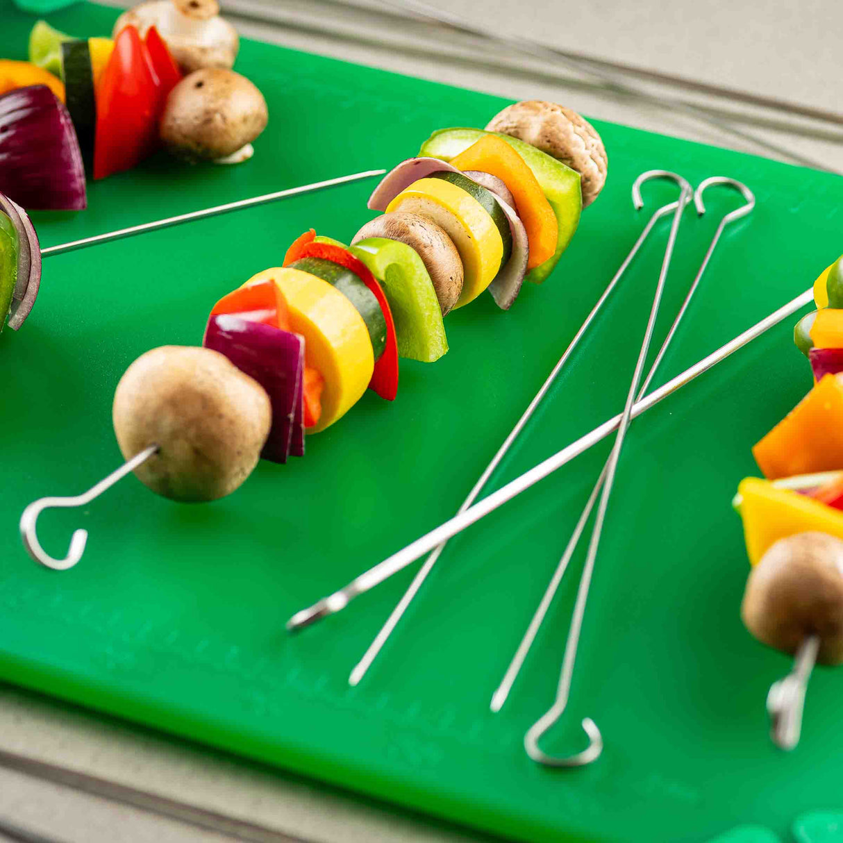 Several stainless steel skewers with raw vegetables—mushrooms, red onion, green, yellow, and red bell peppers—are arranged on a bright green cutting board. Some dishwasher safe skewers are loaded with veggies, while others are empty.