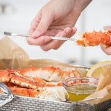A person uses a chrome plated shellfish fork to extract meat from a bright orange king crab leg. The crab legs rest in a metal basket lined with brown paper, next to a lemon wedge and a small bowl of herb-infused melted butter.