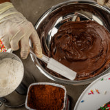 A gloved hand holds a BPA free white spatula, mixing glossy chocolate batter in a metal bowl. Nearby are a container of cocoa powder, a bowl of flour, and colorful sprinkles scattered on a gray surface.