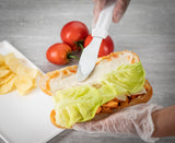 A gloved hand uses a stainless steel sandwich spreader to smooth mayonnaise on a sandwich roll with lettuce and sliced chicken. Fresh tomatoes sit nearby, with a white plate of potato chips on the side, all set on a light gray surface.