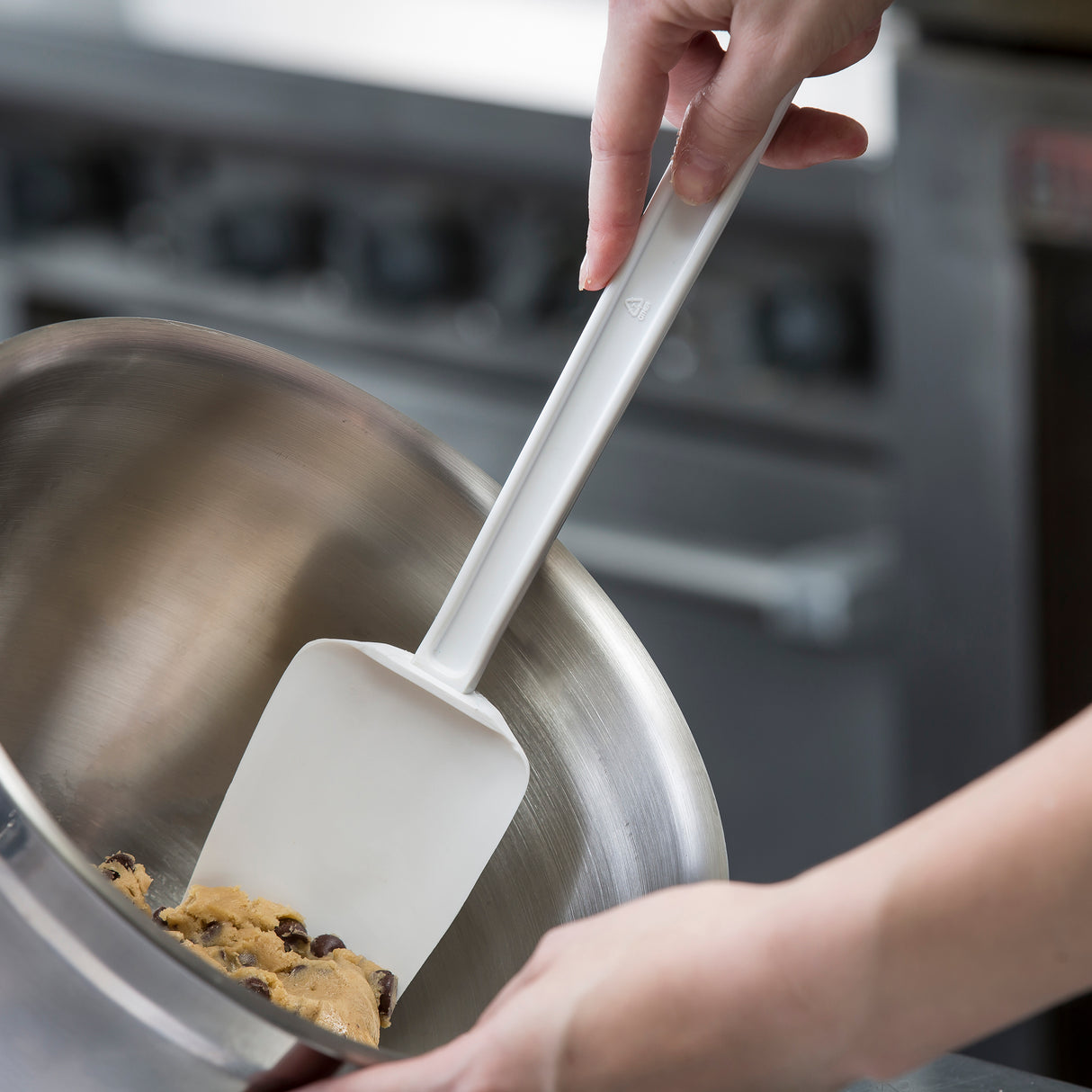 A person uses a heat resistant spatula to scrape chocolate chip cookie dough from the side of a large metal mixing bowl, in a kitchen setting with a stove visible in the background.