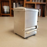 A small, rectangular stainless steel napkin dispenser sits on a countertop. Its shiny surface reflects light, and a stack of napkins is visible through the front opening. The background is softly blurred, showing wooden shelves and kitchen items.