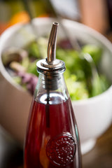 A close-up of a glass bottle with a chrome plated Free Flow Speed Pourer spout, filled with red vinaigrette. In the blurred background, a white bowl holds a green salad with leafy vegetables and serving utensils.