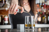 A woman with long brown hair pours whiskey from a bottle fitted with a chrome plated Free Flow Speed Pourer into a glass on a bar counter. A large gray whiskey stone sits nearby, while shelves of colorful liquor bottles blur in the background.