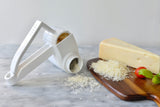 A white cheese grater with a stainless steel drum sits on a marble surface, grated cheese spilling out. Beside it, a wooden cutting board holds a wedge of cheese, fresh basil leaves, cherry tomatoes, and more grated cheese.