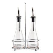 Two glass bottles with stainless steel pourers sit side by side in a silver metal holder with a central handle. This elegant oil and vinegar set features wide bases, narrow necks, and round feet, all displayed against a white background.