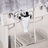 Close-up of a shiny metal beverage dispenser spigot with a lever handle, attached to a clear polycarbonate container. The reflective spout is centered above a removable drip tray with vertical slots.