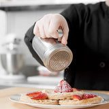 A person in a black sleeve dusts powdered sugar onto waffles topped with sliced strawberries on a plate, using a stainless steel sifter, in a bright kitchen setting.