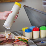 A gloved hand squeezes creamy white dressing from a 16 oz Squeeze Dispenser with a ValveTop onto a sandwich filled with roast beef, cheese, tomato, spinach, and onion. Several condiment bottles with colored lids sit on the counter in the background.