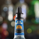 A close-up of a bottle of DeKuyper Blue Curacao liqueur with a black TableCraft Free Flow Pourer. The label is blue with an orange slice graphic. The blurred background features circular light spots, creating a festive atmosphere.