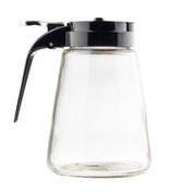 A clear, empty TableCraft Products Modern Glass Dispenser features a black plastic lid and handle. Its trapezoid shape has a wide base and narrower top, with a lever for pouring. Dishwasher safe, it’s set against a plain white background.
