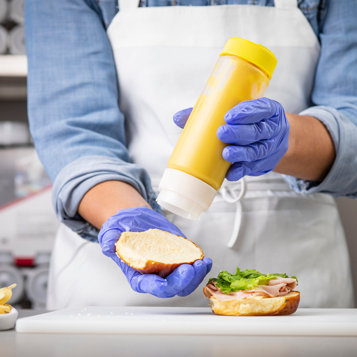 A person wearing blue gloves and a white apron squeezes yellow mustard from an NSF Invertatop Squeeze Bottle End Cap onto a sandwich bun with lettuce and deli meat, in a kitchen or deli setting.