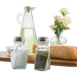 A glass oil bottle, a recycled green tint glass vase with white roses, a loaf of bread, and salt and pepper shakers sit on a wooden board. The scene has a clean, rustic feel with clear containers and an elegant arrangement.