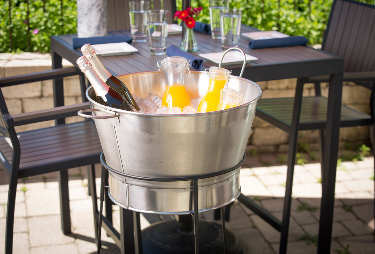 A TableCraft Products stainless steel tub filled with ice holds two bottles of sparkling wine and two carafes of orange juice. The tub sits on a stand beside an outdoor table set with glasses, napkins, and red flowers amid lush greenery.