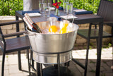 A TableCraft Products stainless steel tub filled with ice holds two bottles of sparkling wine and two carafes of orange juice. The tub sits on a stand beside an outdoor table set with glasses, napkins, and red flowers amid lush greenery.