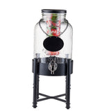 A clear glass TableCraft Industrial Collection metal beverage dispenser with a black spigot stands on a tall black beverage dispenser base. Inside, fruit and herbs float, while a blank round label hangs from a chain beneath the black lid.