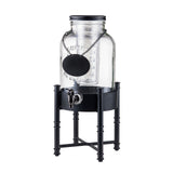 A clear glass TableCraft Industrial Collection metal beverage dispenser with a black lid sits on a tall black metal beverage dispenser base. It features a silver spigot and a black round chalkboard tag for labeling, offering a modern and minimalistic style.