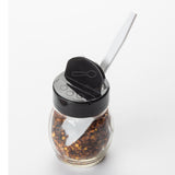 A small glass jar filled with red chili flakes has a black TableCraft flip top plastic lid featuring a pop-up opening for a spoon. A white plastic spoon rests at an angle through the lid, set against a plain white background.