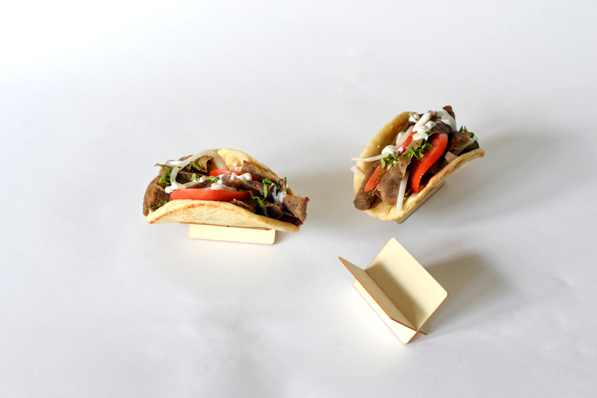 Two tacos filled with sliced meat, lettuce, tomato, onions, and white sauce sit in TableCraft Disposable Taco Taxi holders on a plain white surface. An empty wood-inspired holder lies partially open in the foreground. The background is minimal and uncluttered.