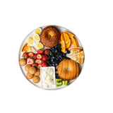 A round melamine serving tray holds bagels, orange slices, hard-boiled eggs, strawberries, blueberries, blackberries, small muffins, cucumbers, cream cheese, and cheesy bread—all neatly arranged on a white background.