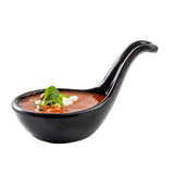 A black melamine soup spoon filled with red tomato soup, topped with a dollop of cream and fresh green herbs, sits on a white background. The long, elegant handle curves upwards to the right, and the spoon is dishwasher safe for easy cleaning.