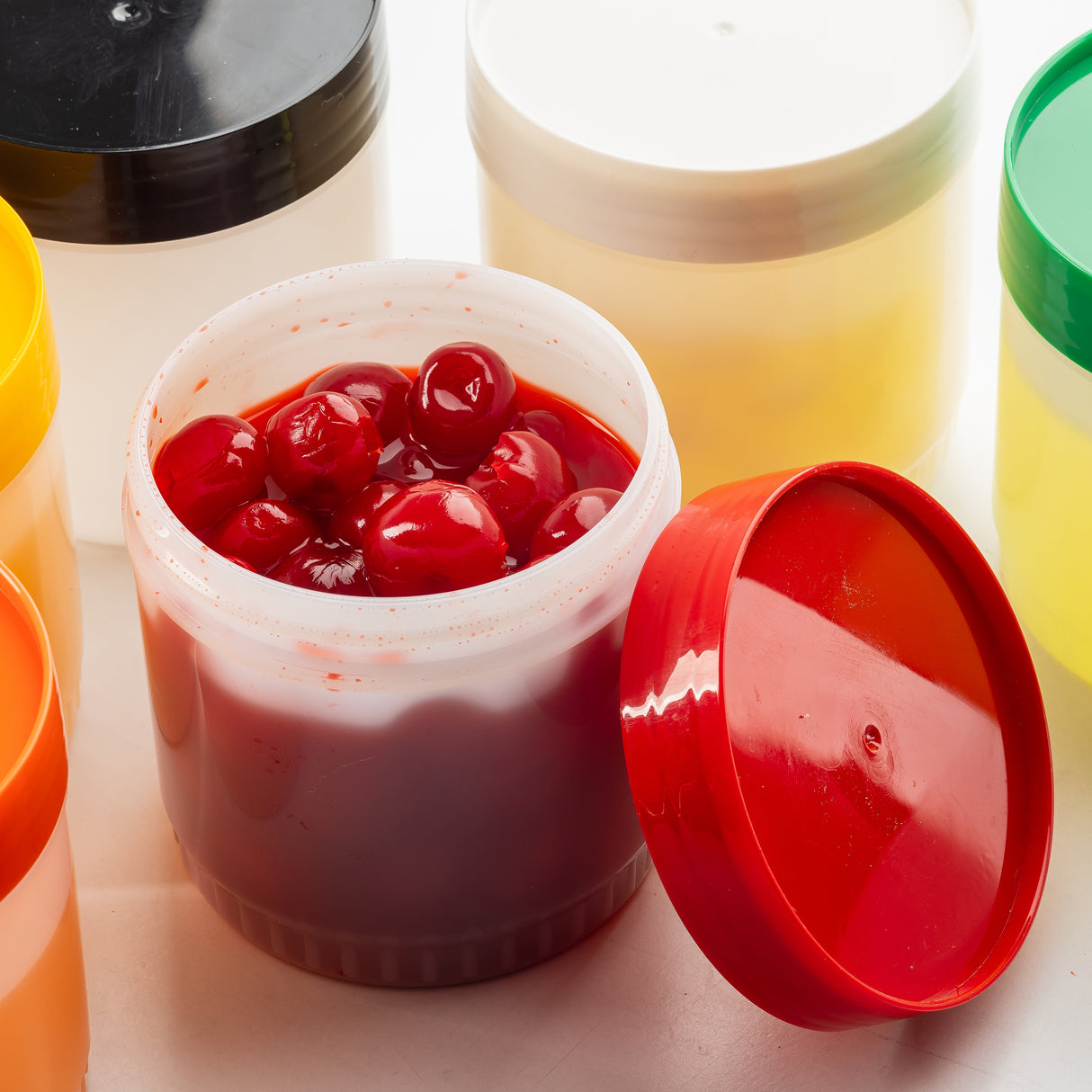 An open TableCraft Products Pint Backup Storage Jar filled with bright red cherries in syrup sits among several closed HDPE storage jars with colorful lids—black, yellow, green, and white—against a white background. The red lid leans nearby.
