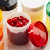 An open TableCraft Products Pint Backup Storage Jar filled with bright red cherries in syrup sits among several closed HDPE storage jars with colorful lids—black, yellow, green, and white—against a white background. The red lid leans nearby.