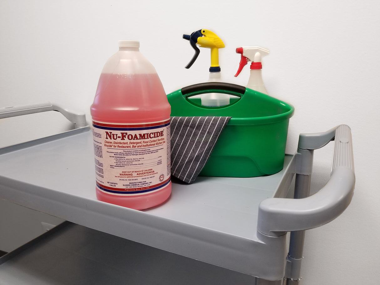 A gray cleaning cart holds a large red jug labeled Glissen Chemical Nu-Foam, an EPA registered all-purpose cleaner, plus a green caddy with two spray bottles and a striped cloth. The background is a plain white wall.