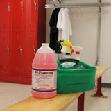 A large bottle of pink Glissen Chemical Nu-Foam all-purpose cleaner sits on a wooden bench next to a green caddy with spray bottles, with red lockers and hanging white shirts in the background, inside a locker room.