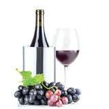 A bottle of red wine sits in a double wall stainless steel wine cooler next to a filled glass. In front, clusters of black and red grapes with a green leaf are artfully arranged, all set against a white background.