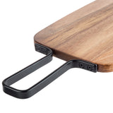 A close-up of a wooden cutting board with a smooth finish and rounded corner. This TableCraft serving paddle features a black metal handle attached with screws, extending outward from one end for easy carrying or hanging.