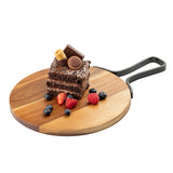 A round acacia serving paddle with a metal handle holds a slice of chocolate cake topped with chocolate pieces and a wafer. Fresh blueberries, raspberries, and a sliced strawberry are arranged around the cake on a white background.