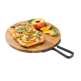A rectangular flatbread topped with peach slices, herbs, and cheese sits on a round acacia serving paddle with a black handle. Fresh peach halves and basil leaves are arranged beside the flatbread against a white background.
