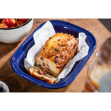 A golden-brown loaf of fruit and nut bread sits in a blue speckle pan lined with parchment paper. One slice is cut, revealing bits of red fruit and nuts inside. A bowl of cherries and a wooden table are visible in the background.