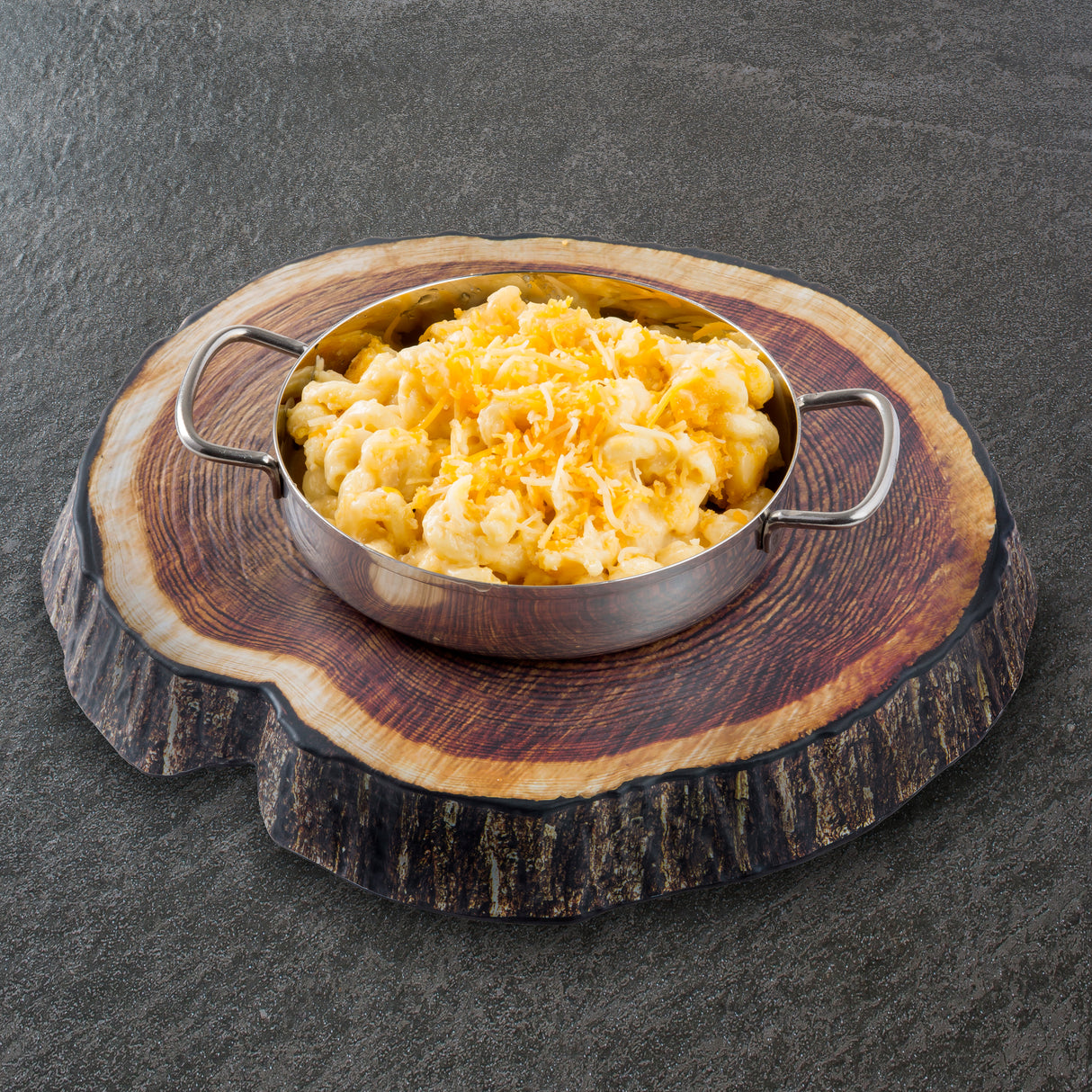 A round metal TableCraft dish with handles is filled with creamy macaroni and cheese, topped with shredded cheese. The dish sits on a platter designed to look like a tree trunk cross-section, placed on a gray textured surface.