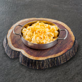 A round metal TableCraft dish with handles is filled with creamy macaroni and cheese, topped with shredded cheese. The dish sits on a platter designed to look like a tree trunk cross-section, placed on a gray textured surface.
