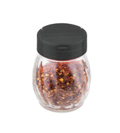A clear glass shaker filled with crushed red pepper flakes, topped with a round black flip top plastic lid, is shown against a plain white background. The pepper flakes are visible through the transparent jar.