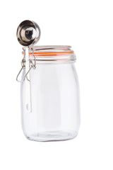 A clear glass canister with an orange rubber seal and metal clip-top lid sits upright. A metal measuring scoop is hooked onto the clasp. The plain white background highlights the canister’s transparency and the scoop’s reflective surface.