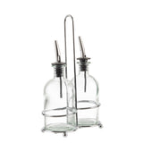 Two clear Oil & Vinegar Bottles with stainless steel pourers sit side by side in a chrome plated rack with a handle. The bottles are held in place by metal rings attached to the holder’s base and midsection, set against a plain white background.