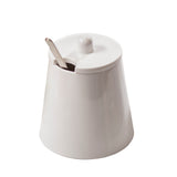 A white ceramic Sierra Collection Sugar Jar with a matching lid featuring a round knob. A metal spoon is partially inserted through a slot in the lid. The bowl has a smooth, glossy finish and is photographed against a plain white background.