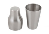 Two stainless steel cocktail shaker pieces are shown side by side on a white background. The left piece is inverted, showing its flat base, while the right stands upright revealing its smooth 20 oz cylindrical interior and brushed metallic finish.