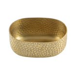 A shallow, oval-shaped aluminum bowl with a textured, hammered surface forming a pebble-like pattern. Smooth rounded edges and a gold powder coat finish softly reflect light across its detailed surface.