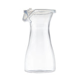 A clear, empty glass carafe with a wide base, narrow neck, and a transparent polypropylene lid. The carafe is shown against a plain white background, and its minimalist design highlights its clean lines and smooth surface.