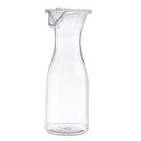 A tall, clear 19 oz polycarbonate carafe with a narrow neck and flared rim is shown upright against a white background. The carafe includes a transparent plastic lid with a metal clasp. Its overall design is simple and modern.
