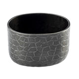 A round, black powder coated aluminum pot with a slightly oval shape. The exterior features a geometric, irregular polygon pattern in relief. The rim is smooth and the inside is unadorned, ideal as a modern sugar packet holder. Background is plain white.