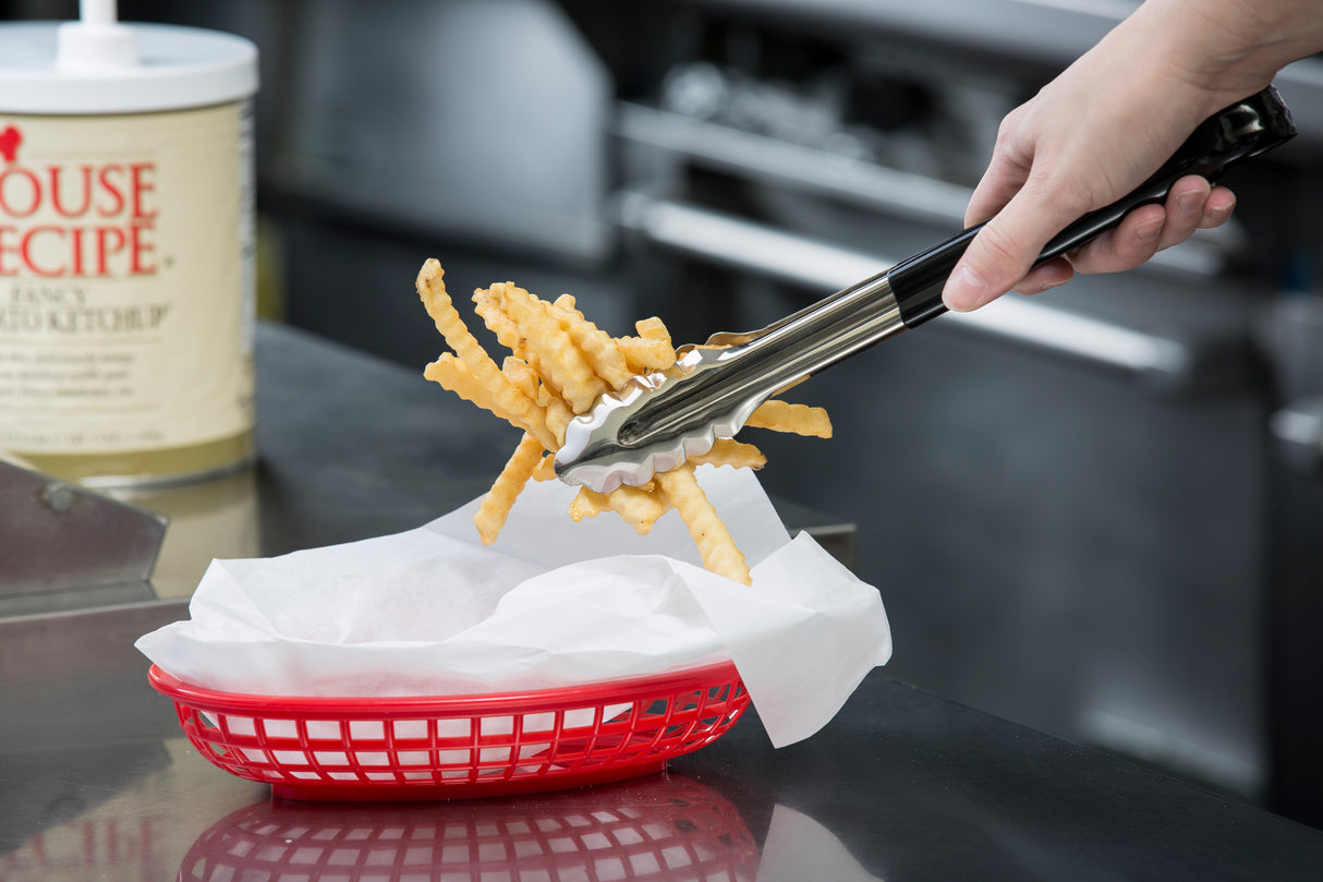 A hand uses dishwasher safe spring steel tongs to place crinkle-cut French fries into a red plastic basket lined with white paper in a commercial kitchen, with a large container of seasoning blurred in the background.