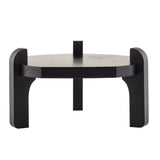 A small, modern, black pinewood stool with a round seat and three thick, curved legs. One leg extends above the seat, adding a sculptural touch. This minimalist piece doubles as a TableCraft beverage dispenser stand on any white background.