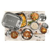A variety of cookware from the Signature Cookware Set, crafted with a sleek stainless steel finish, is filled with colorful dishes—baked casserole, seasoned rice, chili, tortillas, and cheese—artfully arranged on a rustic white wooden surface.